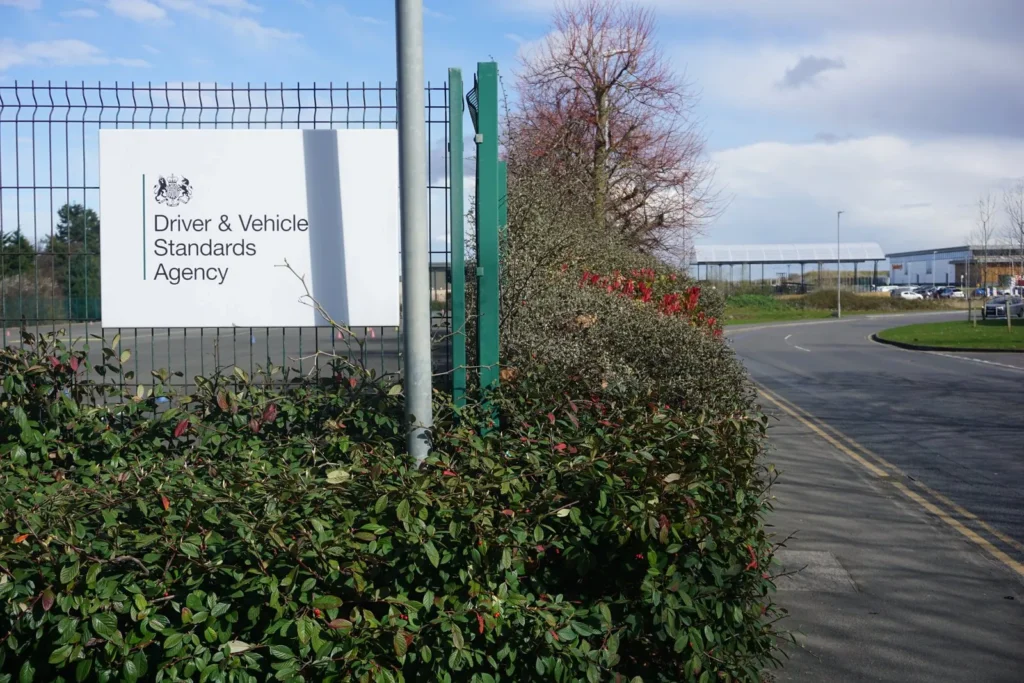 Driver and Vehicle Standards Agency sign on a green mesh fence beside a road curving away