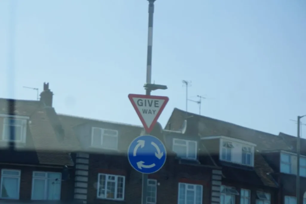 Give way triangle above a blue mini roundabout sign on a pole in front of terraced buildings