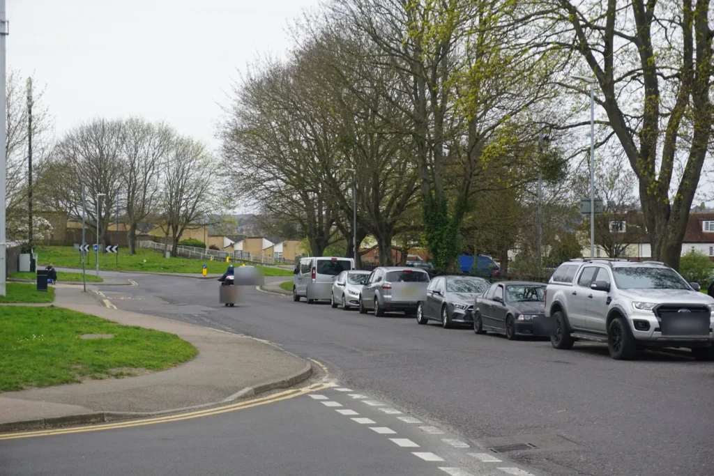Row of cars parked along the kerb beyond a junction, with a scooter rider coming towards the camera
