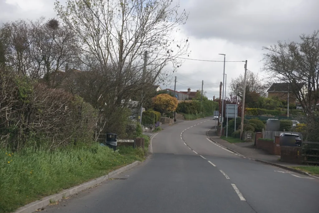 Road dipping and curving into a village, with houses and a parked car on the right