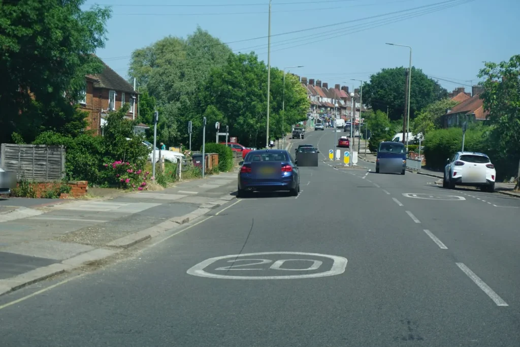 Busy residential road with a painted 20 roundel, parked cars, keep left bollards and traffic ahead