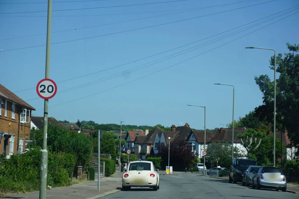 Suburban road with a 20 mph sign, semi detached houses and cars ahead under a blue sky