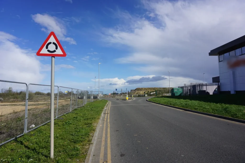 Red triangle roundabout ahead sign beside a road with double yellow lines under a blue sky