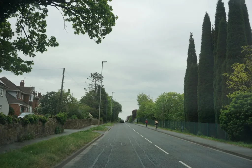 Straight two lane road with a dashed centre line, a low stone wall and houses on the left