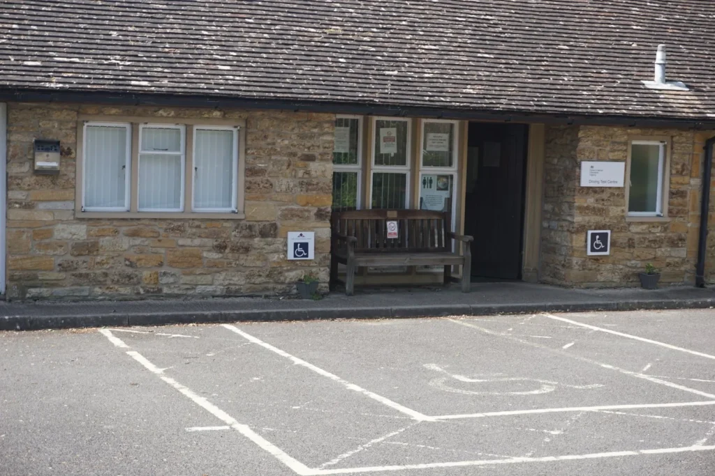 Stone building with a wooden bench by the open door, a Driving Test Centre sign and disabled bays